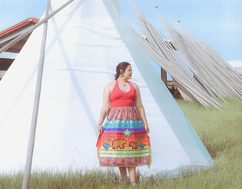 A woman in a traditional red dress standing among white teepees in a field — documentary photograph.